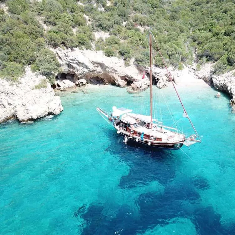 A wooden gulet anchored in a turquoise cove below green hills