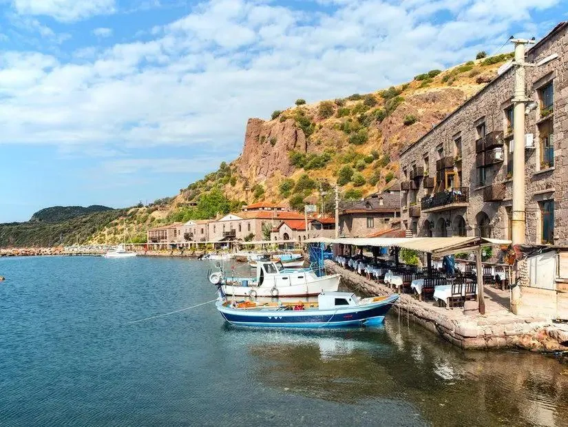 Fishing boats and waterfront restaurant tables in front of the stone buildings of Assos harbour