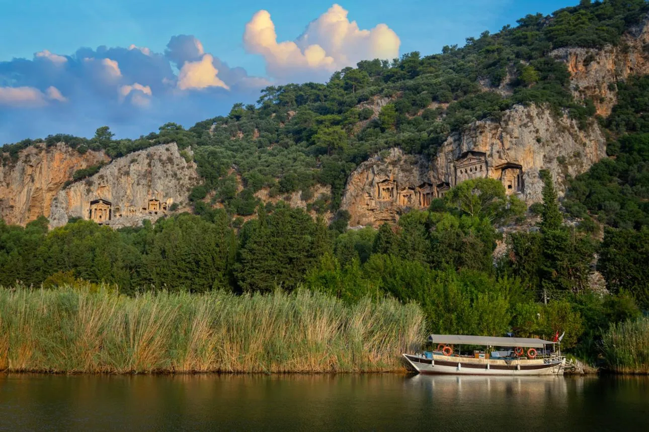 Temple-fronted rock tombs in the cliff above reeds and a river boat
